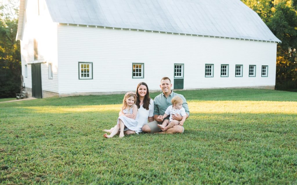 Family of four sitting on grass in front of white barn | Family Photographers Montgomery Alabama | Melissa Sheridan Photography | summer session