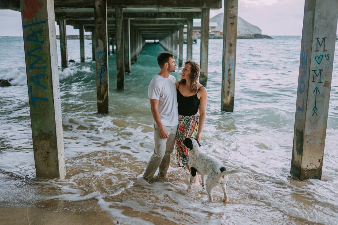 man and woman at beach under a dock with dog | beach engagement photos