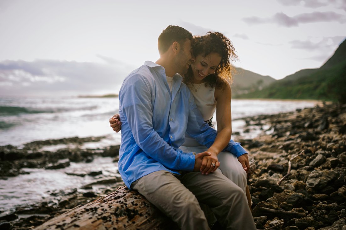 man and woman laughing sitting on log on beach | beach engagement photos