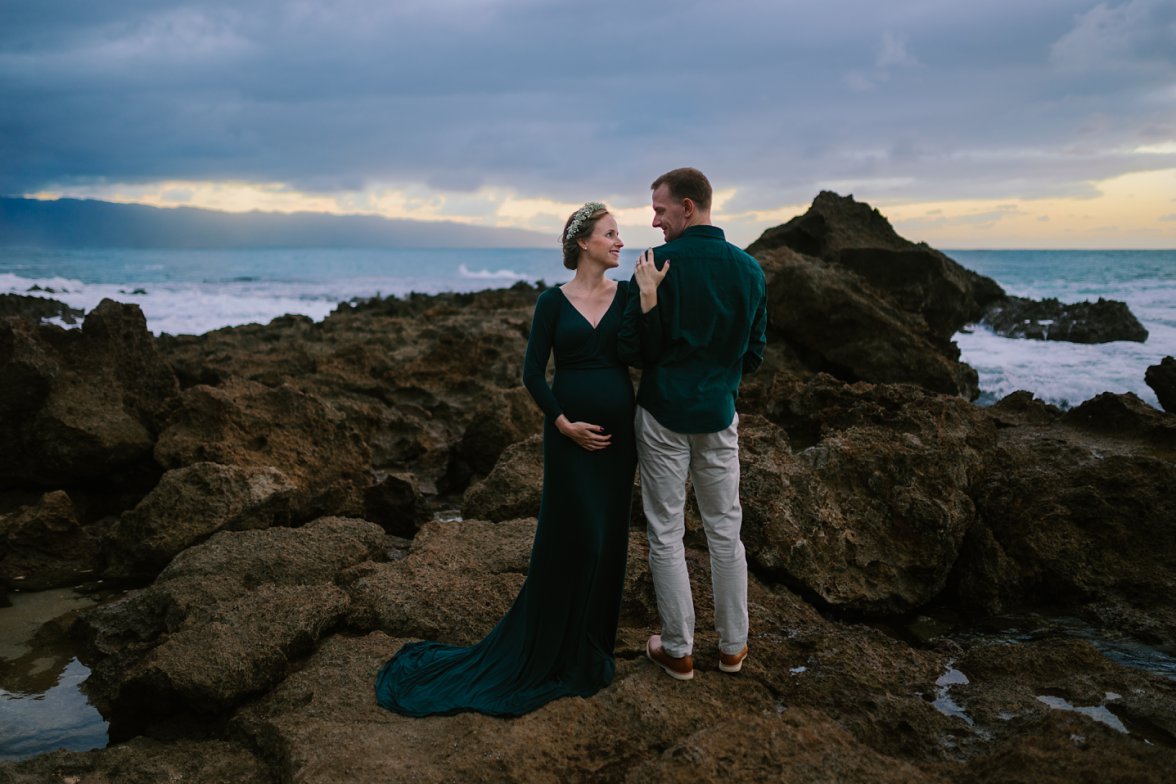 couple expecting baby standing on rocks with ocean behind them 