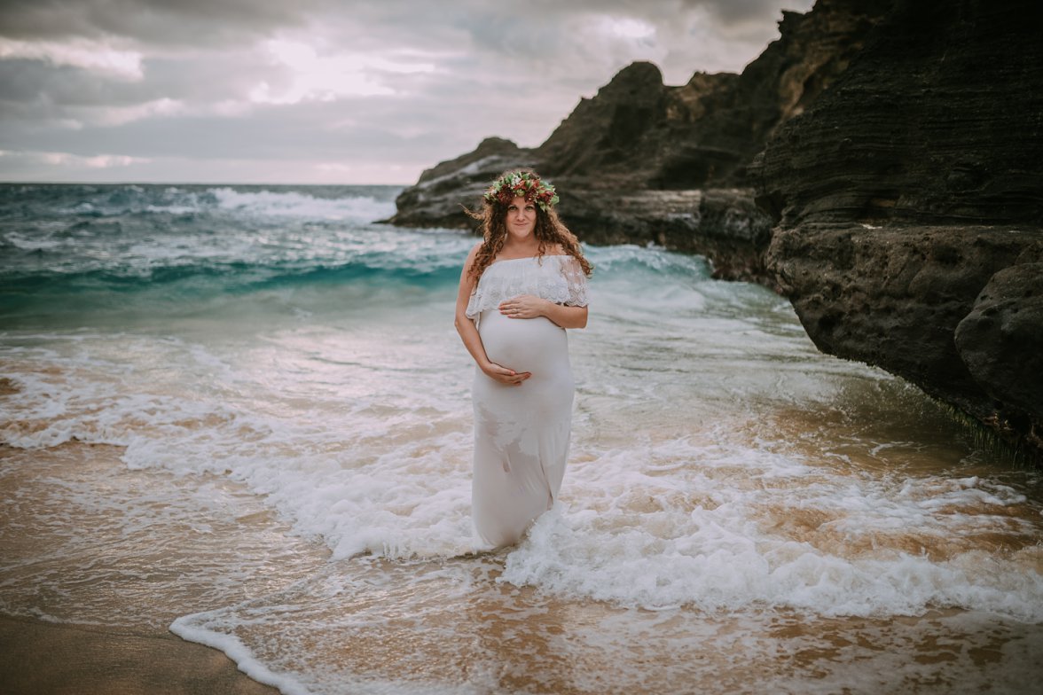 pregnant woman in white dress standing in ocean by rocks 